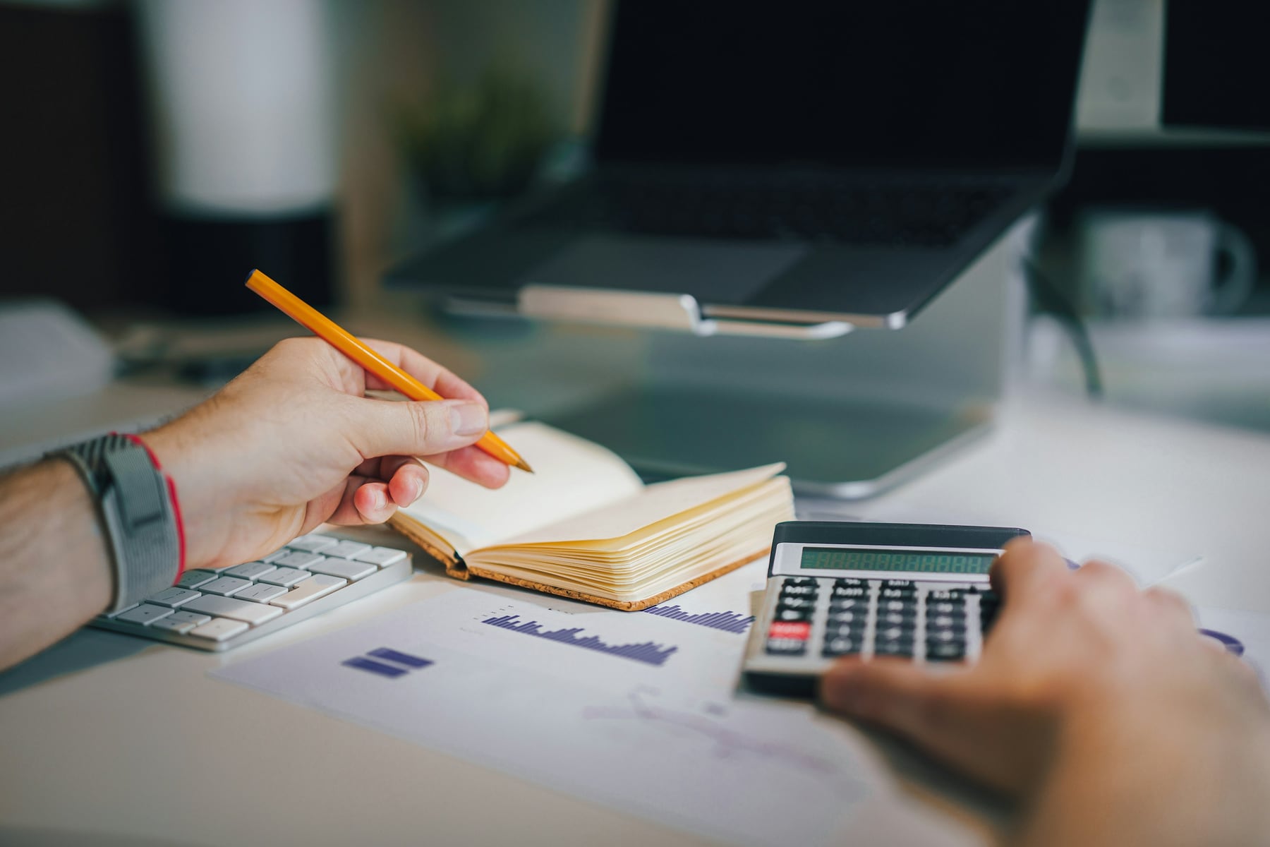 A person calculating a study budget with a calculator, laptop and notebook