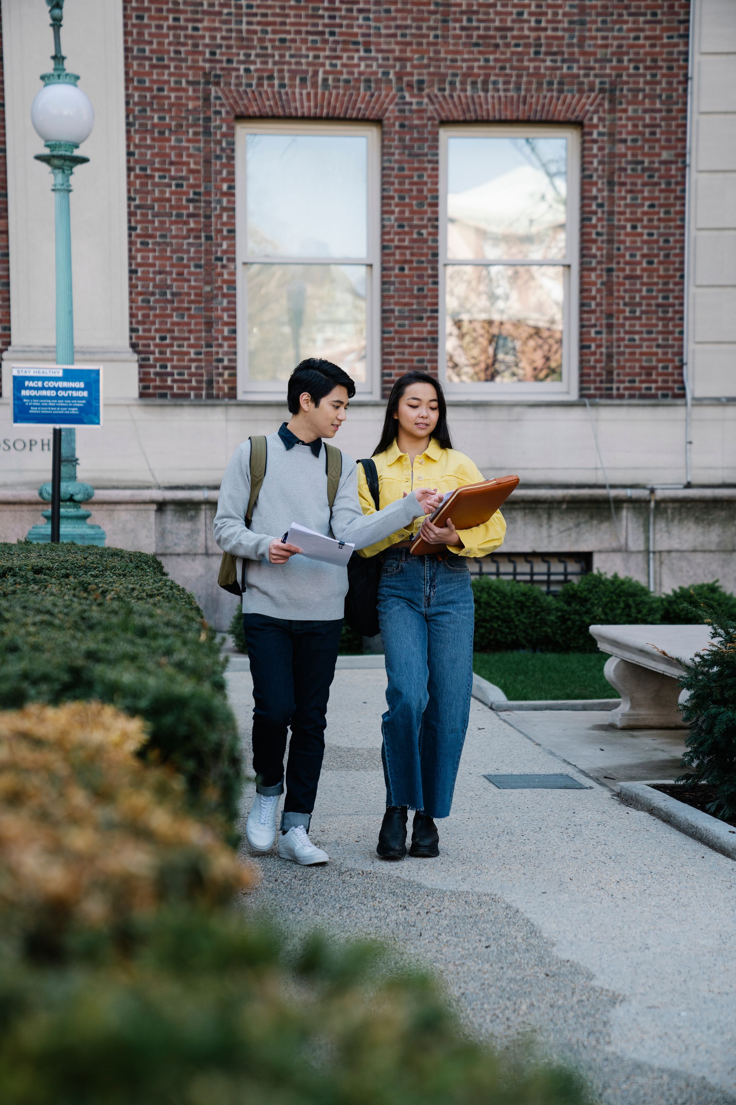 Two students carrying documents walk through a university campus and discuss admission options