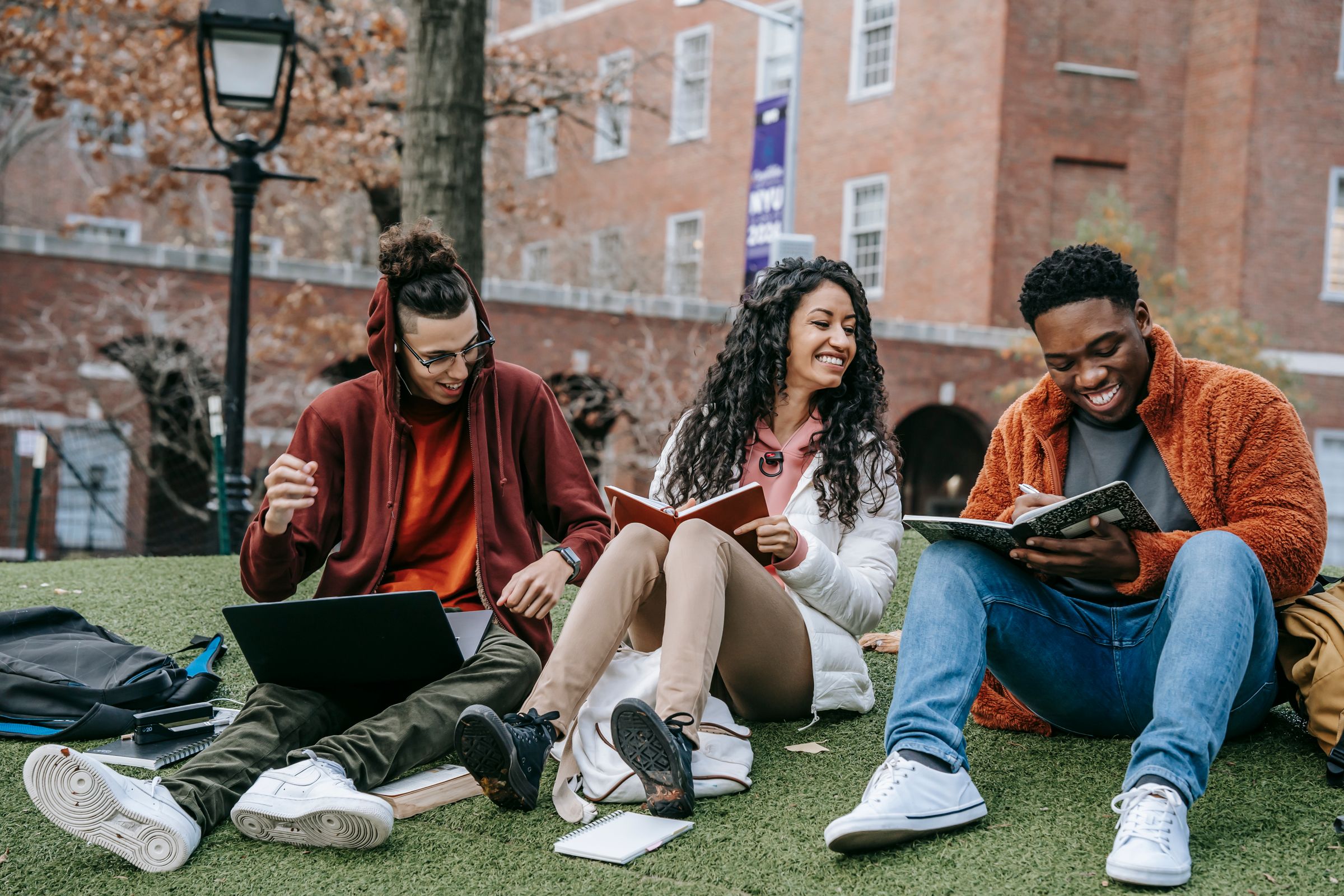 Three university students study together outdoors on campus with a laptop and notebooks
