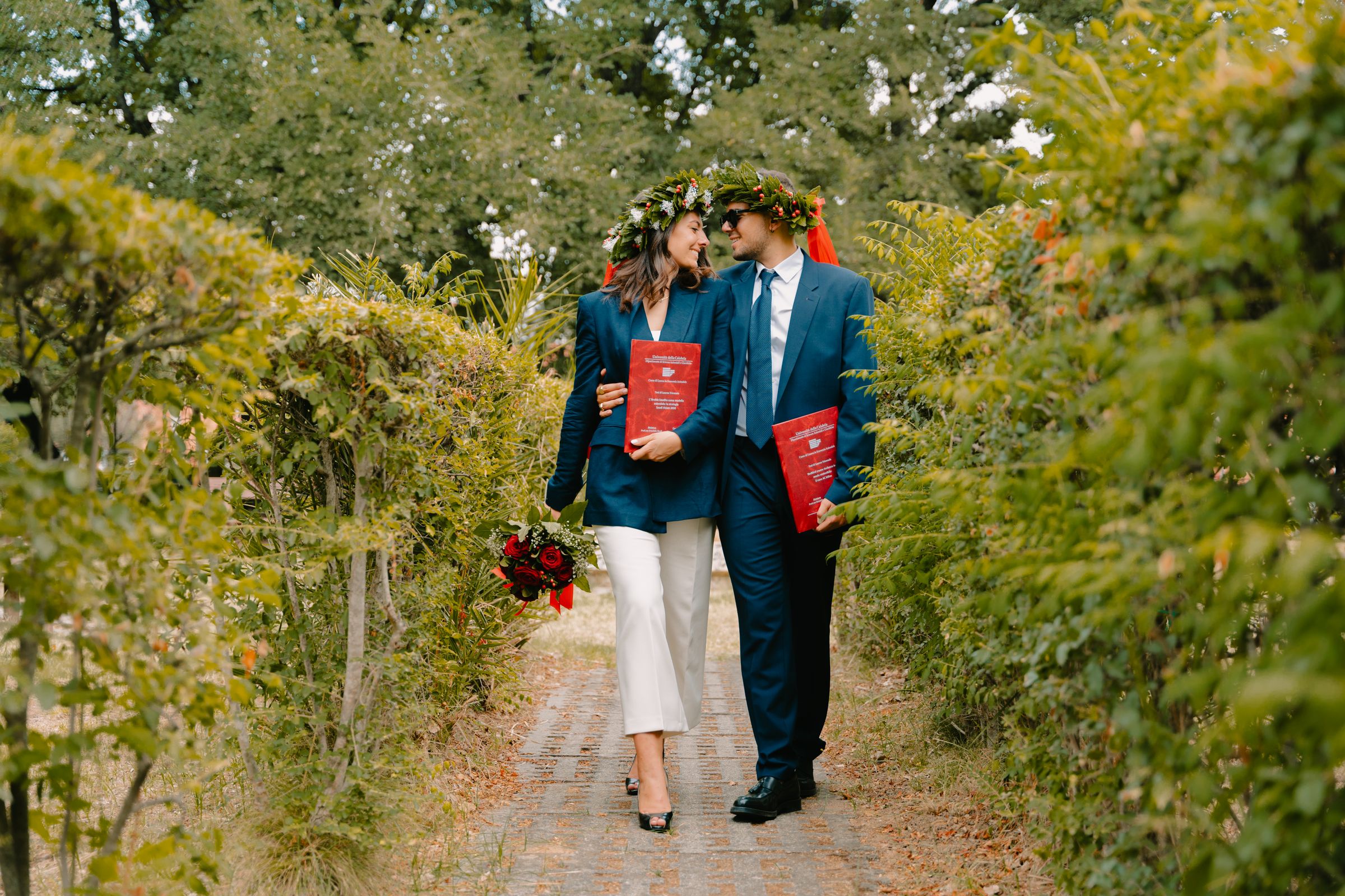 Two university graduates wearing laurel wreaths walk outdoors in Italy